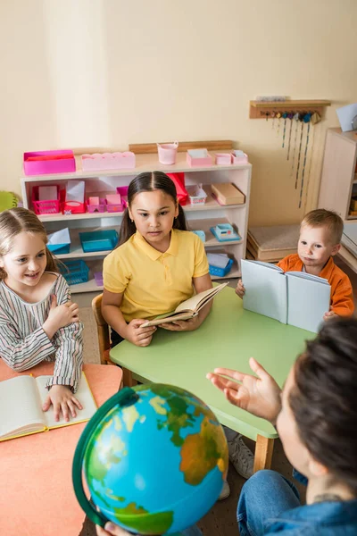 Enseignant pointant avec la main près du globe devant les enfants interracial — Photo de stock