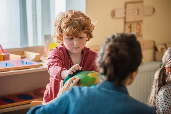 Кудрявый мальчик смотрит на глобус рядом с размытыми учительницей и девочкой в школе Монтессори — стоковое фото