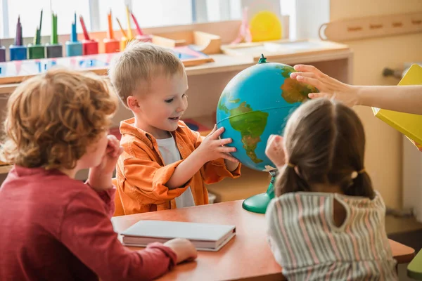 Garçon étonné regardant globe près flou enseignant et les enfants à l'école montessori — Photo de stock