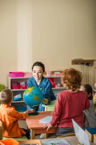 Profesor sonriente hablando con niños cerca del mundo en el aula - foto de stock