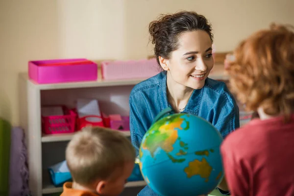 Vista posteriore di ragazzi sfocati vicino al globo e insegnante sorridente nella scuola montessori — Foto stock