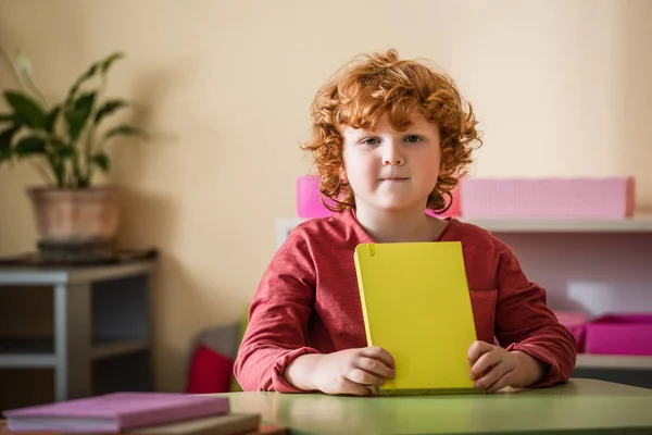 Rousse garçon regardant caméra tandis que assis au bureau avec le livre — Photo de stock