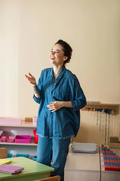 Professor alegre em camisa jeans rindo com os olhos fechados na escola montessori — Fotografia de Stock