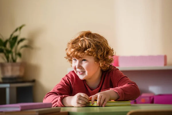 Menino feliz, encaracolado sorrindo perto de livros borrados na escola montessori — Fotografia de Stock