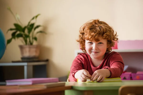 Pelirroja y rizado chico sonriendo en cámara cerca borrosa libros en montessori escuela - foto de stock