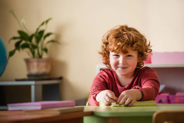 Ruiva e menino encaracolado sorrindo para a câmera perto de livros borrados na escola montessori — Fotografia de Stock