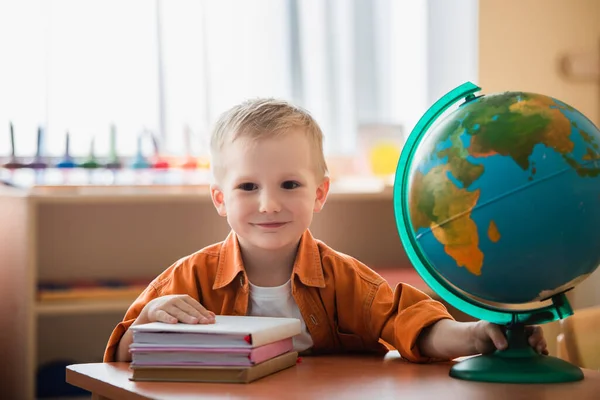 Cheerful boy looking at camera near books and globe on desk — Stock Photo