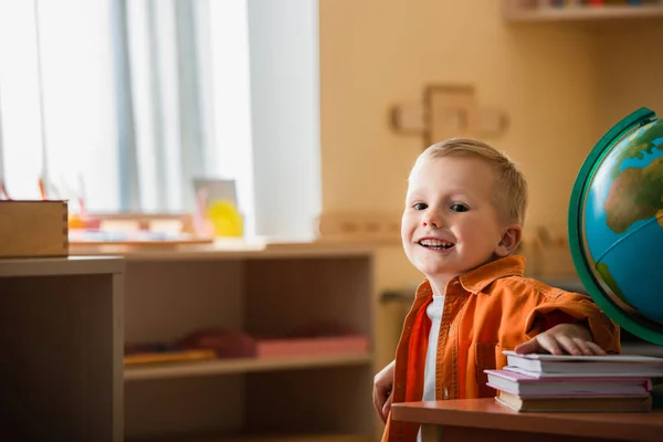 Niño feliz sonriendo a la cámara cerca del globo y libros en la escuela montessori - foto de stock