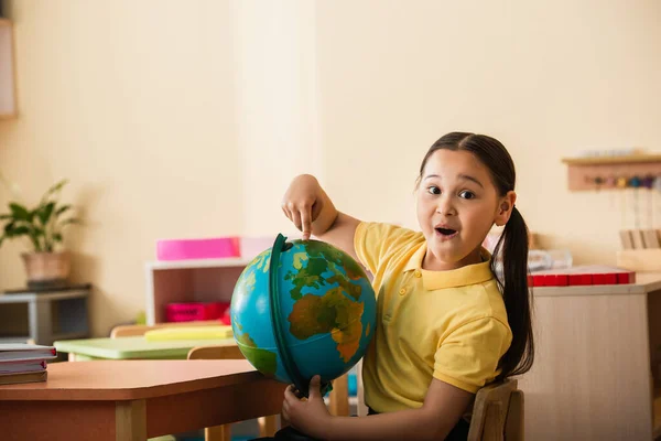 Erstaunt asiatisches Mädchen zeigt auf Globus und blickt in Montessori-Schule in die Kamera — Stockfoto