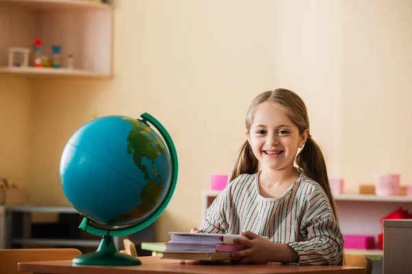 Happy kid sitting with books near globe in montessori school — Stock Photo