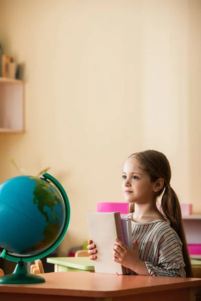 Girl holding books while sitting near globe in montessori school — Stock Photo