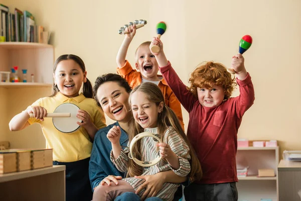 Niños multiétnicos emocionados tocando instrumentos musicales cerca del profesor feliz - foto de stock