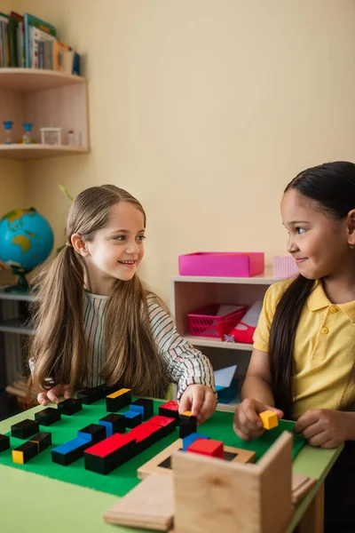 Happy interracial girls looking at each other while playing with wooden blocks in montessori school — Stock Photo