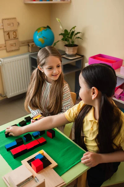 Menina feliz olhando para amigo asiático jogando jogo de cubos multicoloridos na escola montessori — Fotografia de Stock