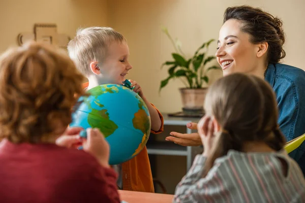 Щасливий хлопчик тримає глобус біля веселого вчителя дітей на розмитому фоні — стокове фото