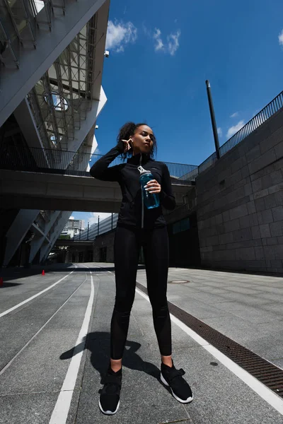 Visão de baixo ângulo da esportista afro-americana em fone de ouvido segurando garrafa de esportes na rua urbana — Fotografia de Stock