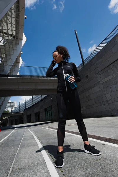Low angle view of african american sportswoman with sports bottle using earphone on urban street — Stock Photo