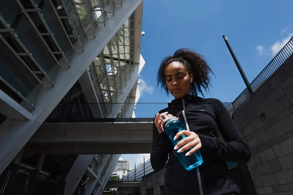 Low angle view of african american sportswoman looking at sports bottle on urban street — Stock Photo