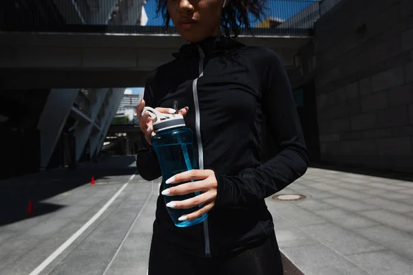 Vue recadrée d'une sportive afro-américaine dans un écouteur tenant une bouteille de sport à l'extérieur — Photo de stock