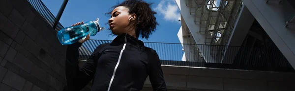 Vue à angle bas de la sportive afro-américaine dans un écouteur tenant une bouteille de sport dans une rue urbaine, bannière — Photo de stock