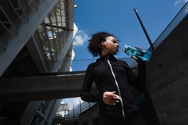 Vista lateral de la deportista afroamericana en auricular sosteniendo botella de deportes al aire libre, vista de ángulo bajo - foto de stock