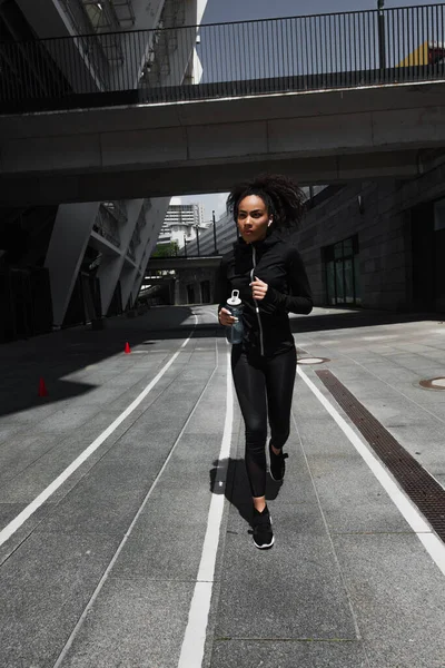 Desportista afro-americana em fone de ouvido segurando garrafa de esportes enquanto corre na rua urbana — Fotografia de Stock