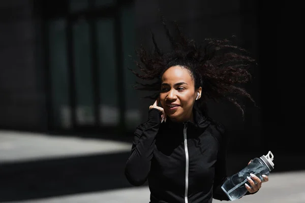 Sonriente corredor afroamericano en auriculares sosteniendo botella deportiva al aire libre - foto de stock