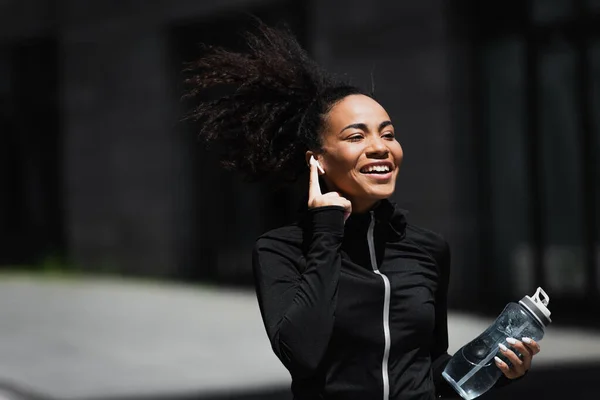 Positive african american jogger with sports bottle using earphone outdoors — Stock Photo