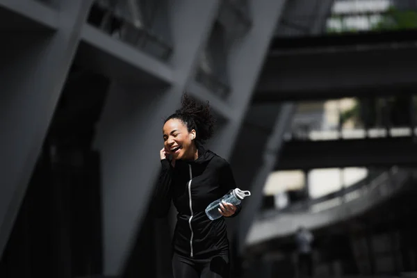 Desportista afro-americana feliz em fone de ouvido segurando garrafa de esportes na rua urbana — Fotografia de Stock
