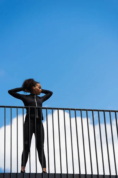 Low angle view of african american sportswoman in earphone standing with closed eyes on bridge — Stock Photo