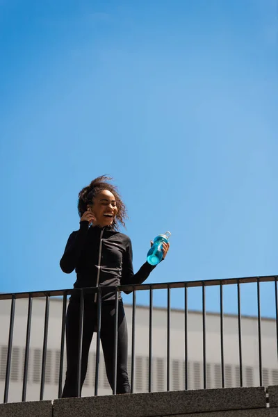 Vista de bajo ángulo de la deportista afroamericana escuchando música en auriculares y sosteniendo la botella de deportes en el puente - foto de stock