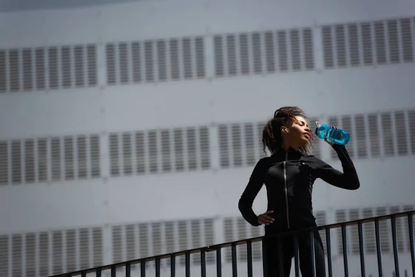 Low angle view of african american sportswoman in earphone drinking water on urban street — Stock Photo