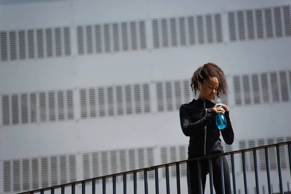 African american sportswoman in earphone holding sports bottle on bridge on urban street — Stock Photo