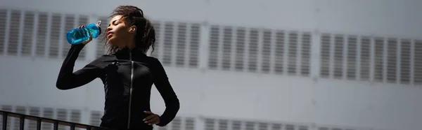 Низкий угол зрения на африканскую американскую спортсменку в наушниках питьевой воды на открытом воздухе, баннер — стоковое фото