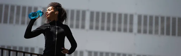 Low angle view of african american sportswoman in earphone drinking water outdoors, banner — Stock Photo