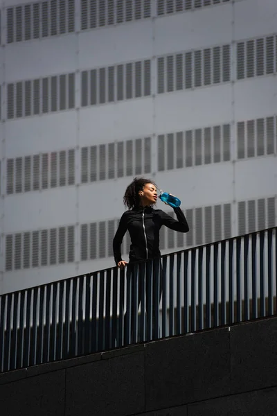 Deportista afroamericana bebiendo agua en puente en calle urbana - foto de stock