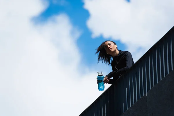Tiefansicht einer afrikanisch-amerikanischen Sportlerin, die eine Sportflasche mit Himmel im Hintergrund hält — Stockfoto