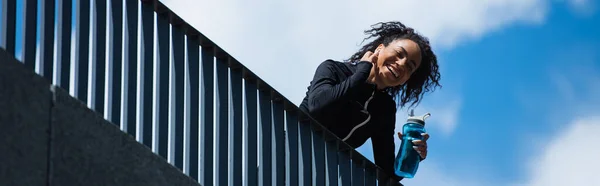 Low angle view of cheerful african american sportswoman in earphone holding sports bottle with sky at background, banner — Stock Photo