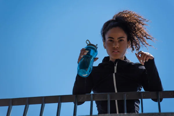 Афроамериканська спортсменка з низьким кутом зору в навушниках, що тримає спортивну пляшку на мосту і дивиться на камеру. — стокове фото