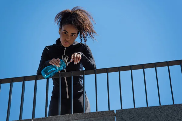 Tiefansicht einer jungen afrikanisch-amerikanischen Sportlerin mit Kopfhörer, die eine Sportflasche mit blauem Himmel im Hintergrund hält — Stockfoto