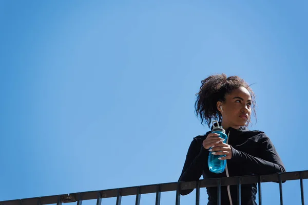 Tiefansicht einer afrikanisch-amerikanischen Sportlerin im Kopfhörer, die eine Sportflasche mit blauem Himmel im Hintergrund hält — Stockfoto