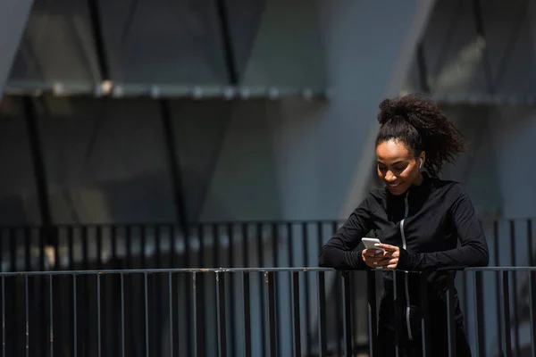Sonriente deportista afroamericana en auricular usando teléfono celular en la calle urbana - foto de stock