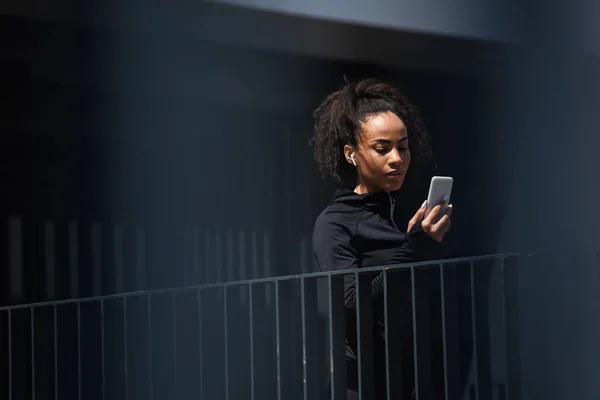 Young african american sportswoman in earphone using mobile phone outdoors — Stock Photo