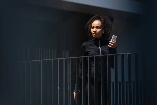 Bastante afroamericana deportista en auricular celebración de teléfono inteligente al aire libre - foto de stock