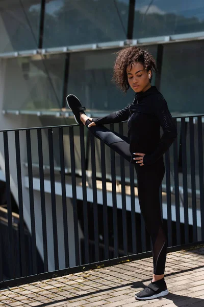 Pretty african american sportswoman in wireless earphone stretching leg on urban street — Stock Photo