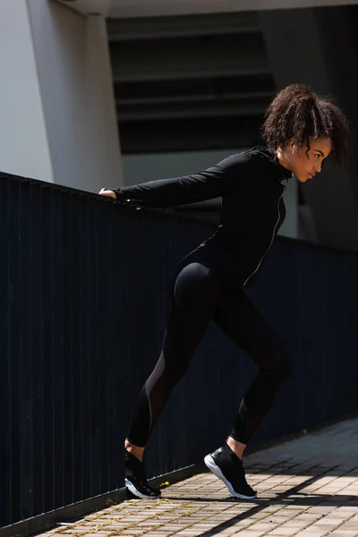 Vista lateral da mulher esportista afro-americana em fone de ouvido aquecendo perto da ponte — Fotografia de Stock