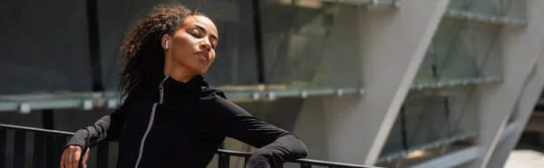 Femme afro-américaine en vêtements de sport et écouteurs debout les yeux fermés près du pont, bannière — Photo de stock