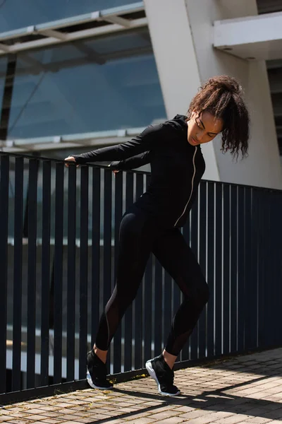 Fit african american sportswoman in earphone warming up beside bridge outdoors — Stock Photo