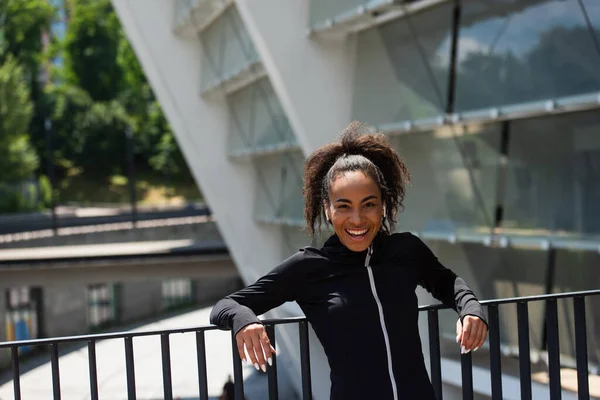 Smiling african american sportswoman in earphones standing near bridge on urban street — Stock Photo