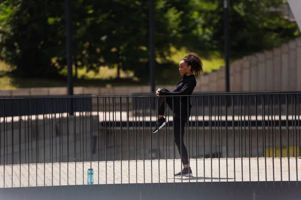 Seitenansicht der fitten afrikanisch-amerikanischen Sportlerin, die Beine in der Nähe von Sportflasche auf Brücke streckt — Stockfoto